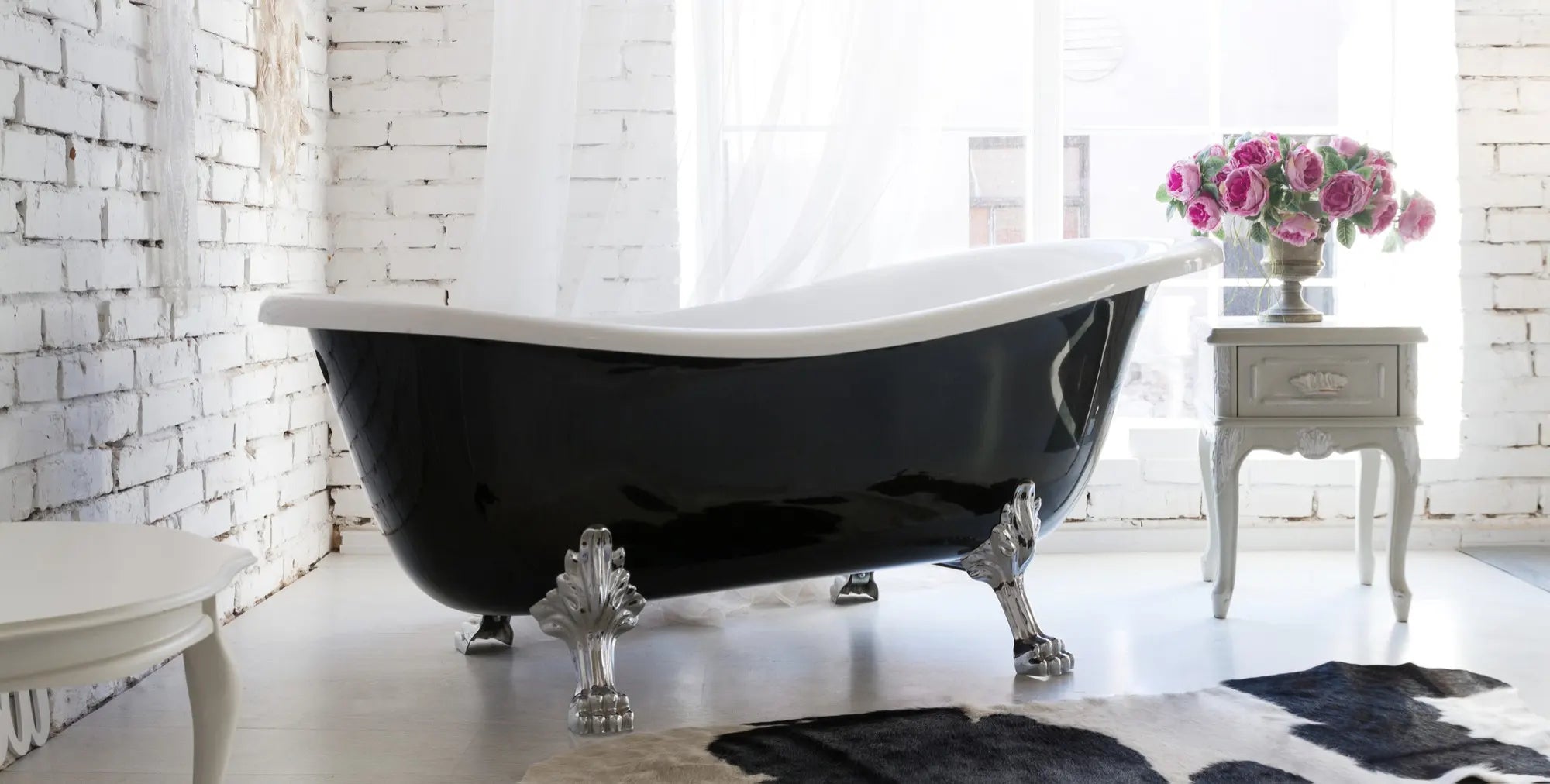Dreamwerks vintage black and white clawfoot bathtub with ornate silver feet, set in a bright, rustic room with white brick walls.