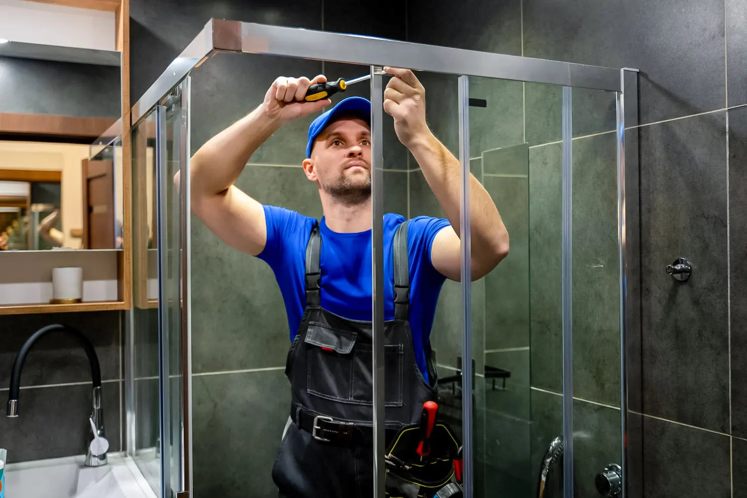 An installer in a blue t-shirt, blue cap, and black work overalls uses a screwdriver to secure the top frame of a modern, chrome-trimmed glass shower enclosure.