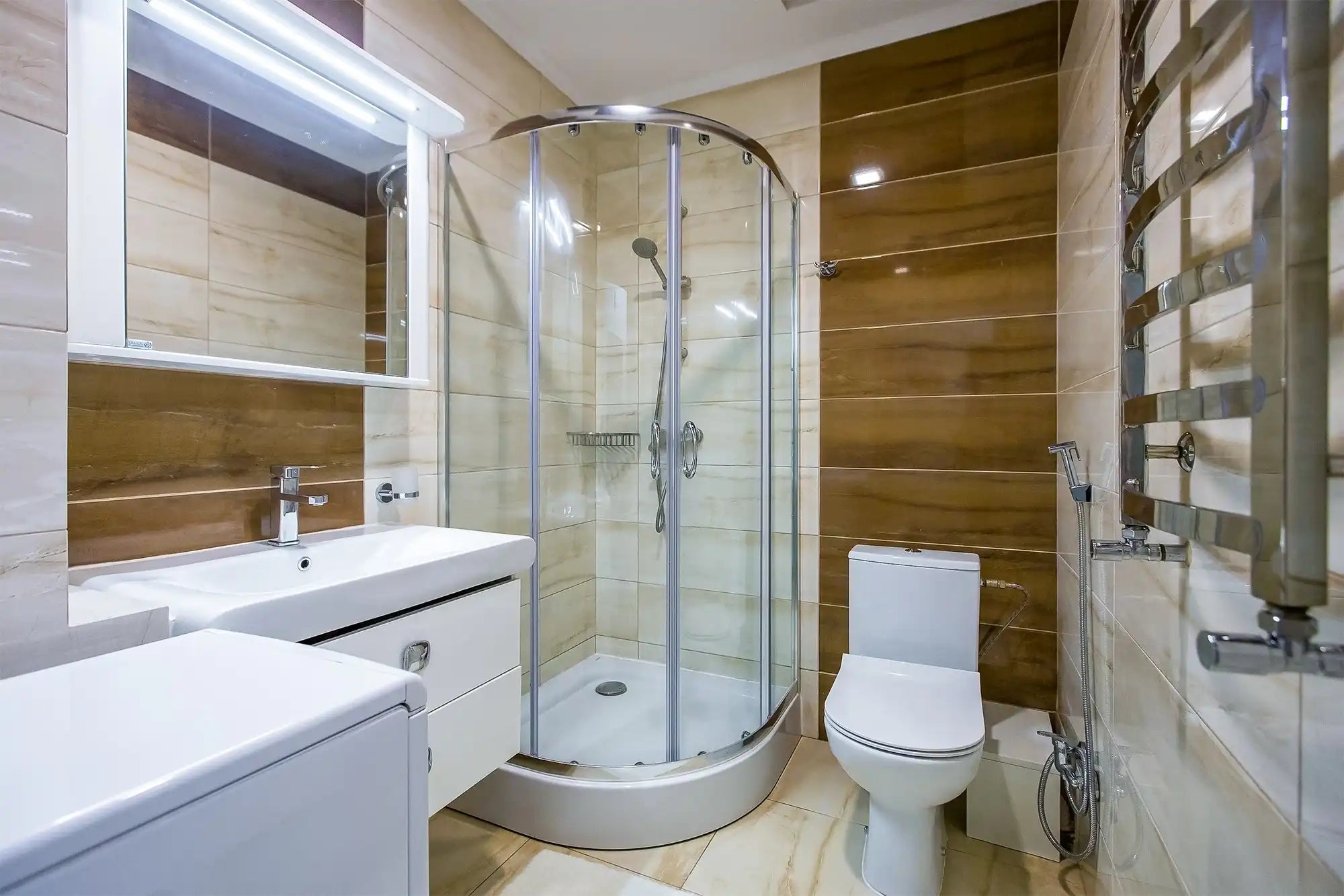 Bright, residential bathroom with brown and beige tiles, featuring a curved glass corner shower kit, a white toilet, a towel warmer rack, and a modern vanity.