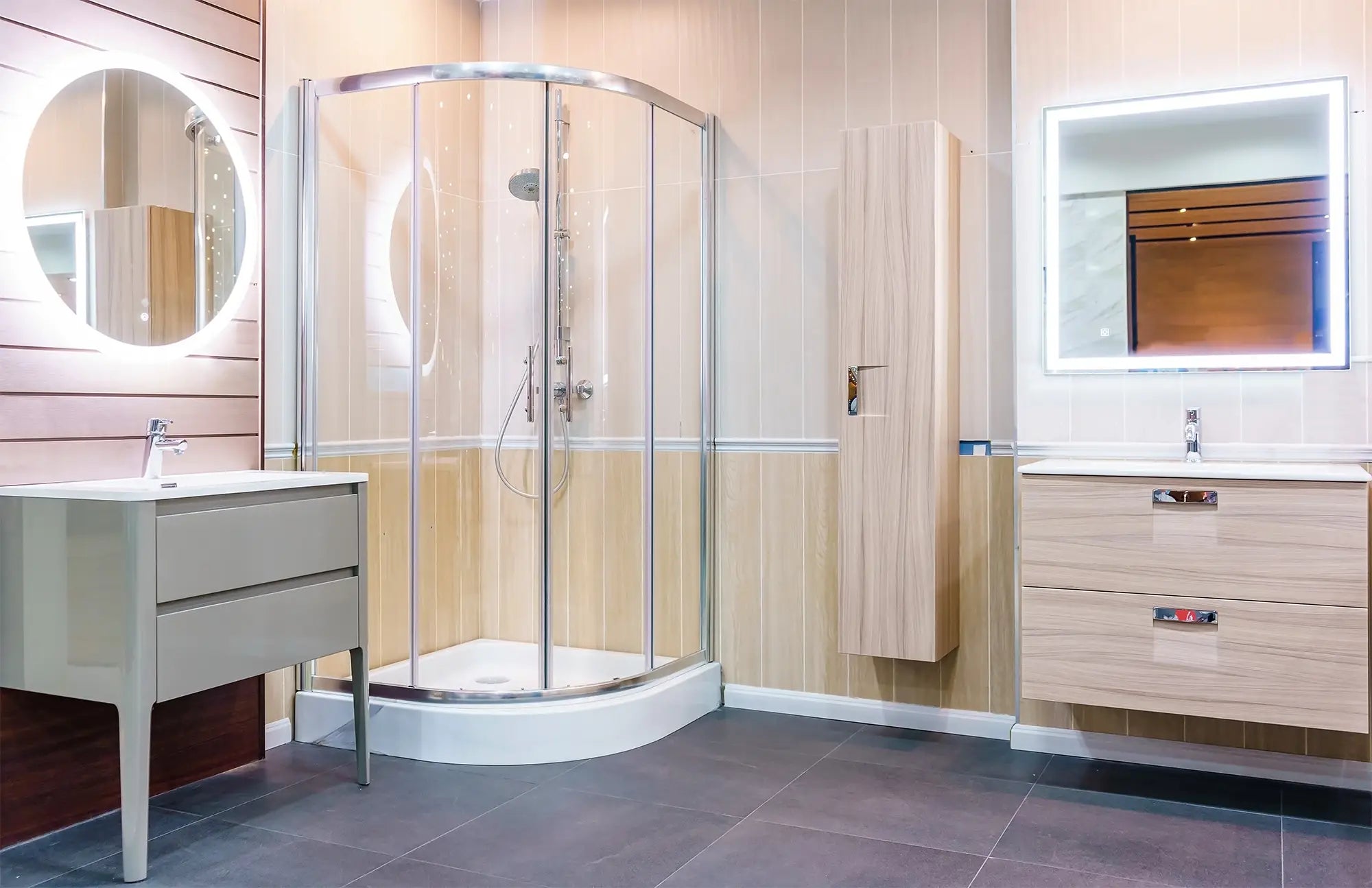 Bathroom showroom display with light wood-tone walls, dark floor tiles, a curved glass corner shower enclosure, and two separate vanities with lighted mirrors.
