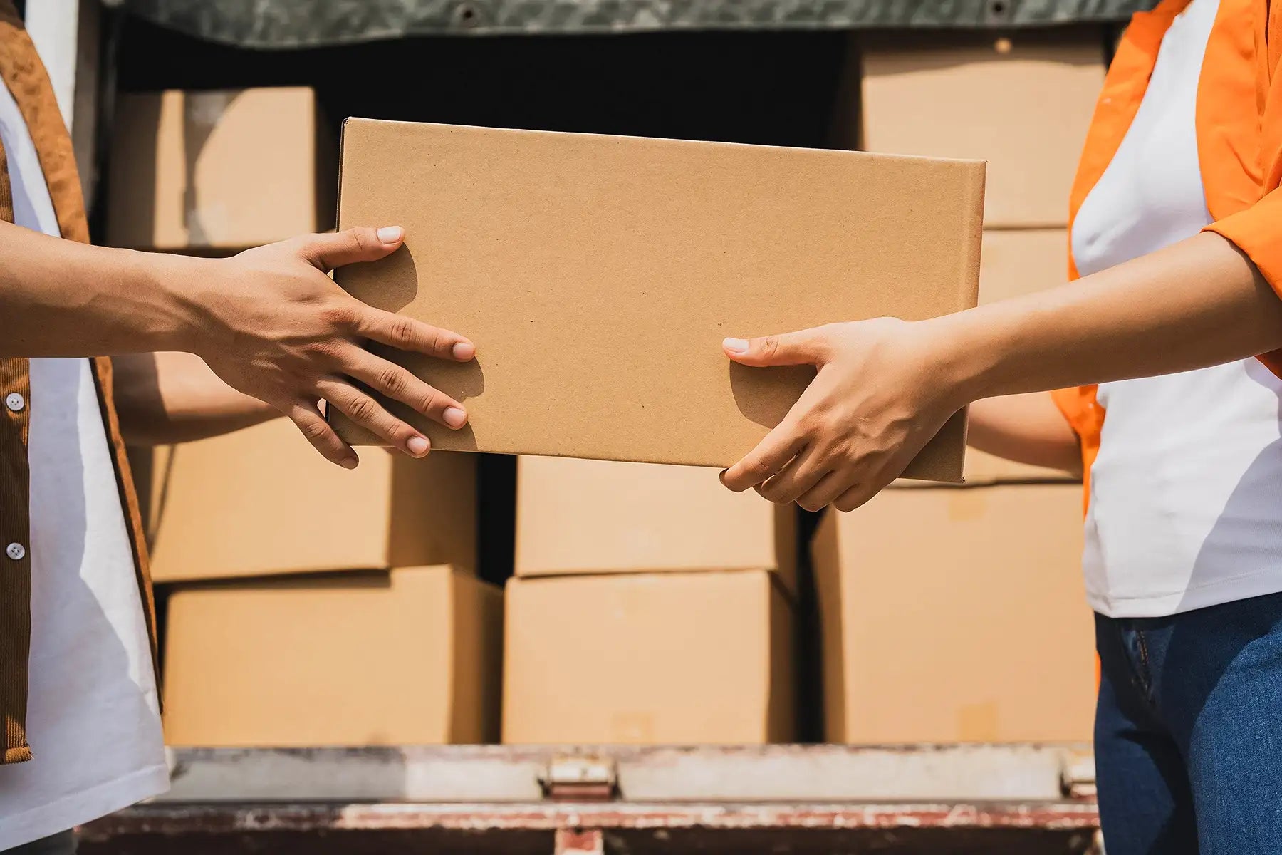 Two people exchanging a cardboard box with stacked boxes in the background