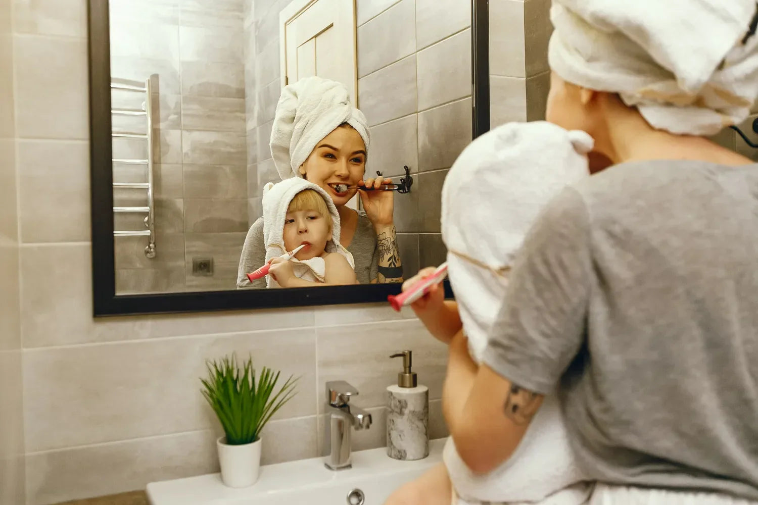 A warm, candid reflection in a black-framed bathroom mirror shows a woman and a young child, both with white towels wrapped around their heads, brushing their teeth together.