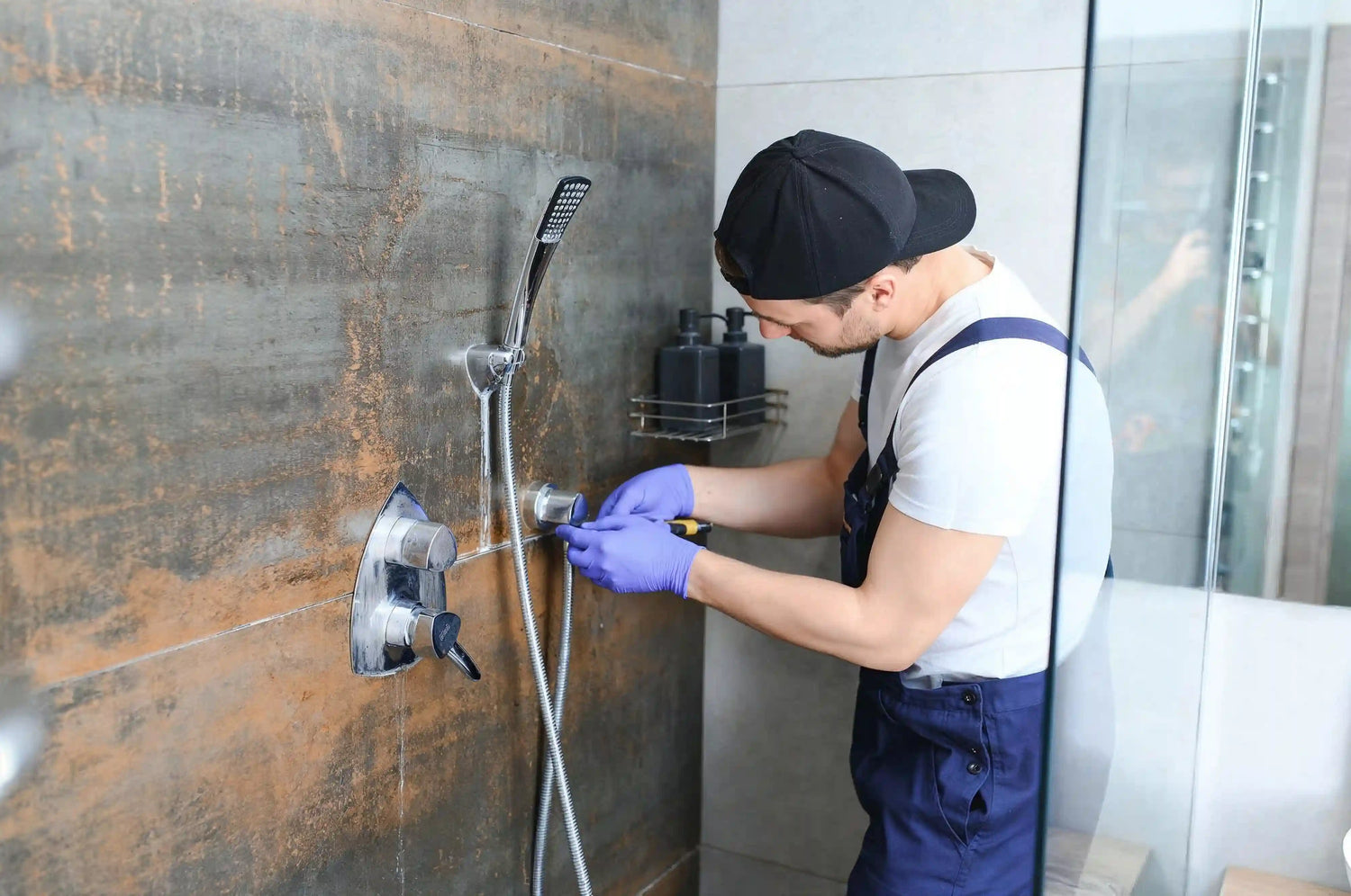 A close-up of a construction worker or installer wearing a tool belt and blue jeans, using a drill to secure hardware into the wall near a modern shower door frame during installation or repair.