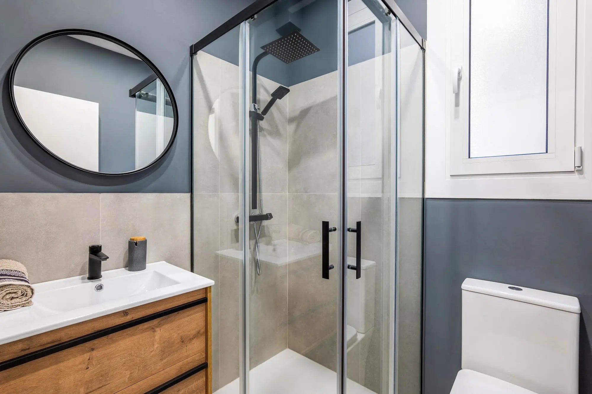 Modern bathroom with black-framed corner shower enclosure, round mirror, and wood vanity.