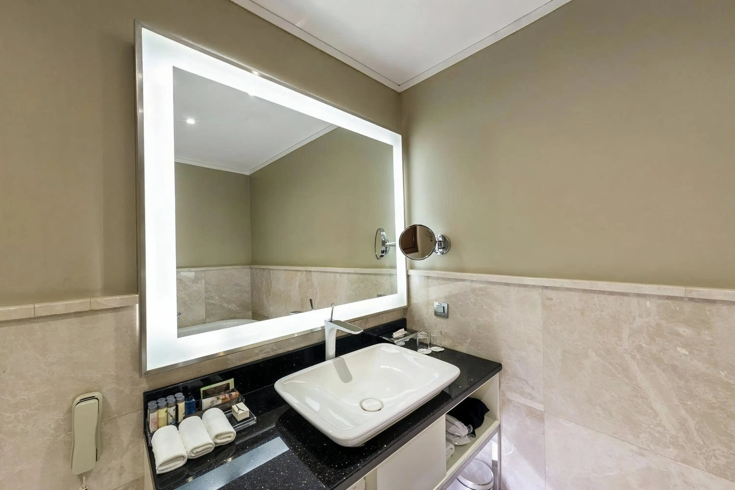 Modern bathroom featuring a large rectangular LED backlit mirror above a white vessel sink with a sleek chrome faucet. The vanity has a black granite countertop, beige tiled walls, and soft ambient lighting, creating a clean and contemporary design.