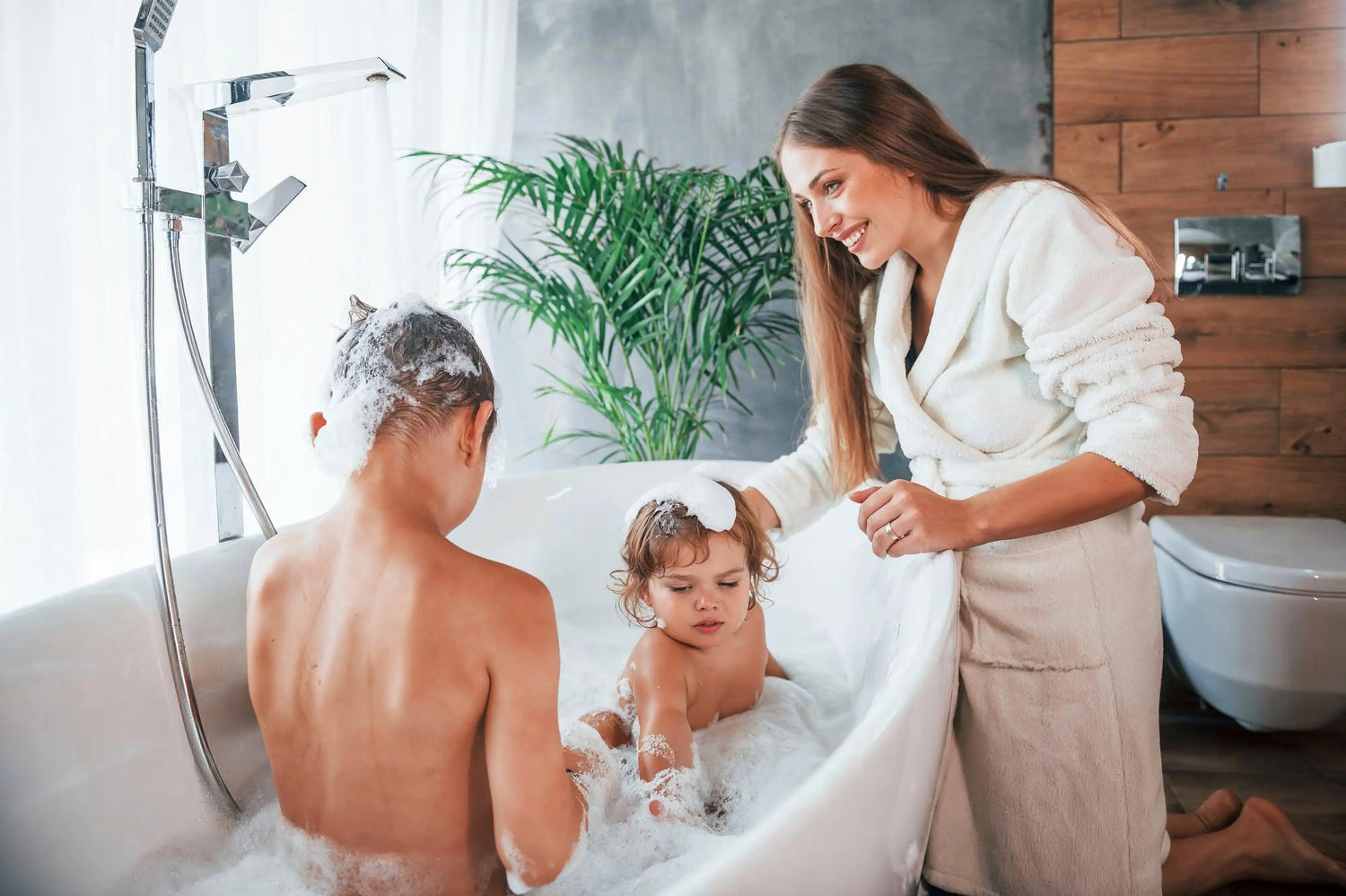 A smiling woman in a white bathrobe watches two children playing in a modern white freestanding bathtub filled with bubbles.