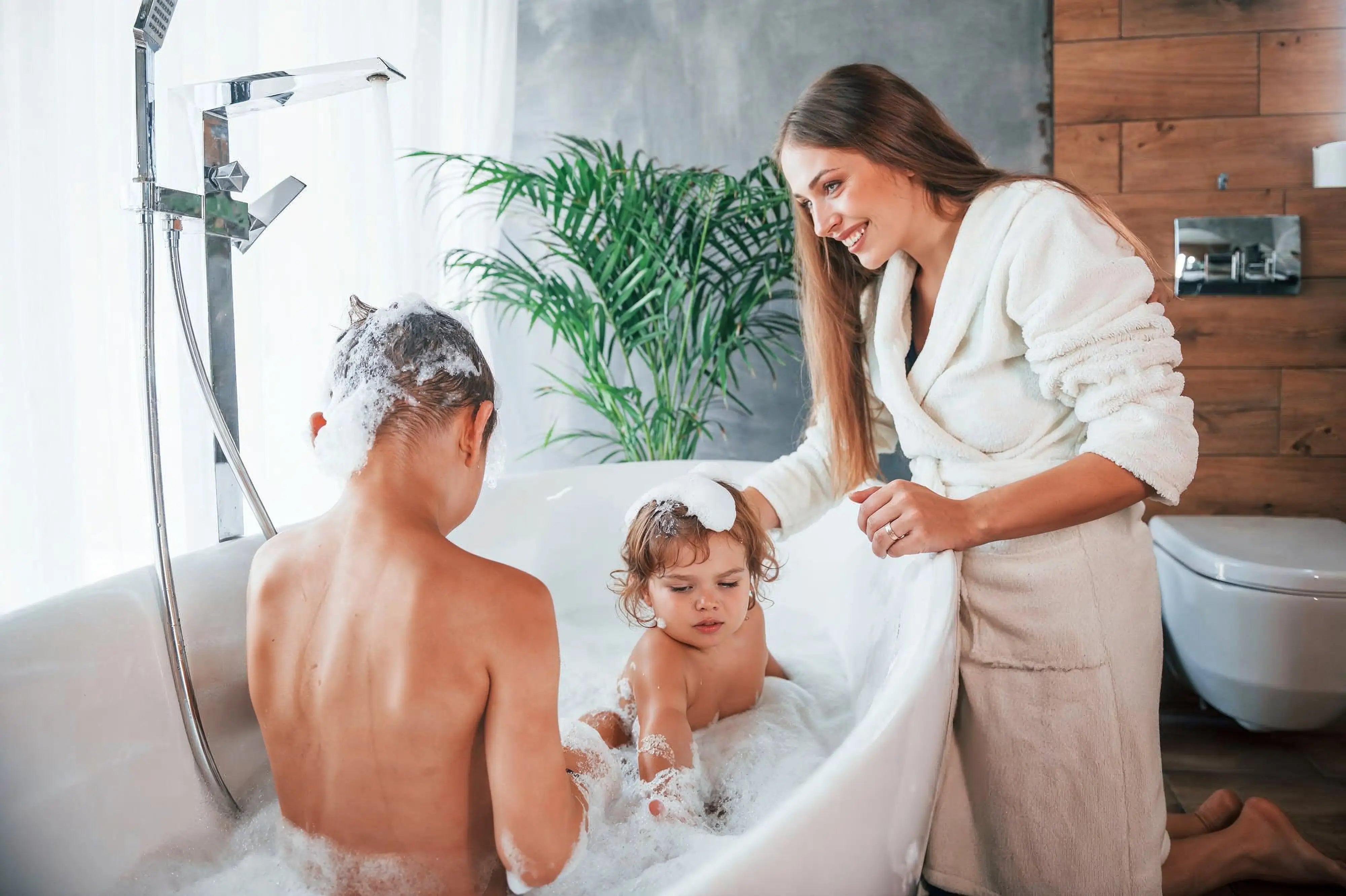 A smiling woman in a white bathrobe watches two children playing in a modern white freestanding bathtub filled with bubbles.