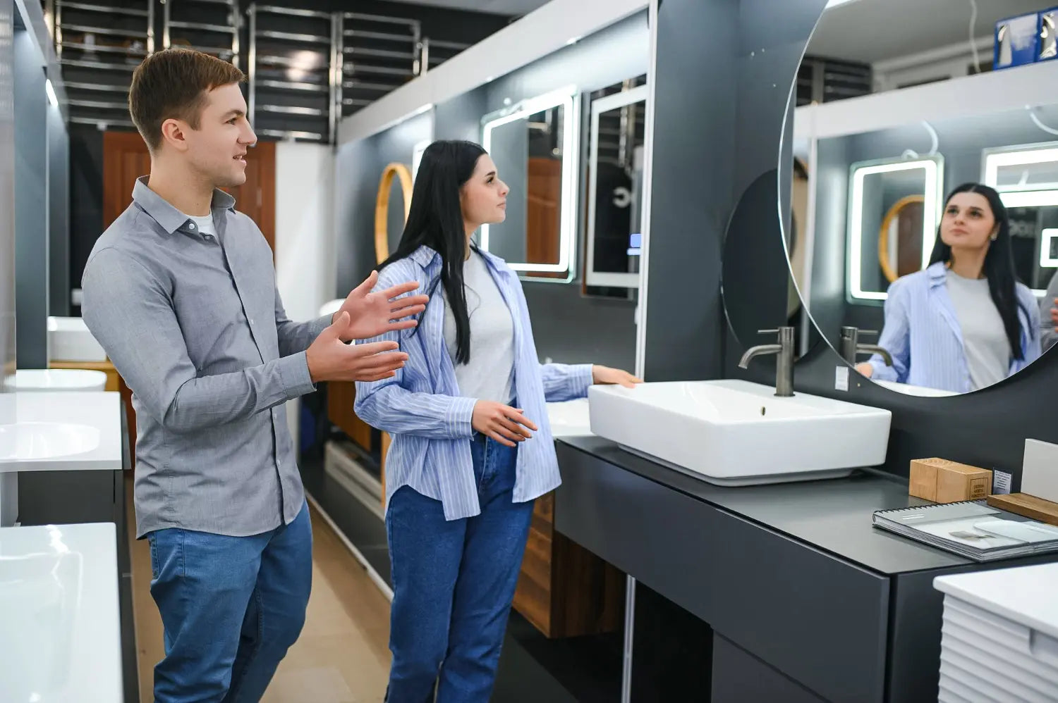 A frameless oval LED mirror with a glowing backlight mounted on white marble-veined wall tiles. Below, a modern white ceramic sink features a chrome faucet and patterned soap dispensers.