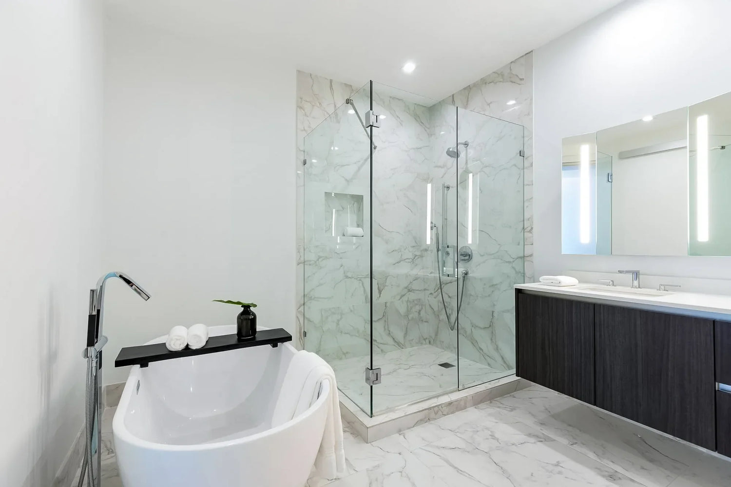 Contemporary bathroom with marble walls, frameless glass shower enclosure, freestanding soaking tub, and a floating double-sink vanity with LED mirrors.