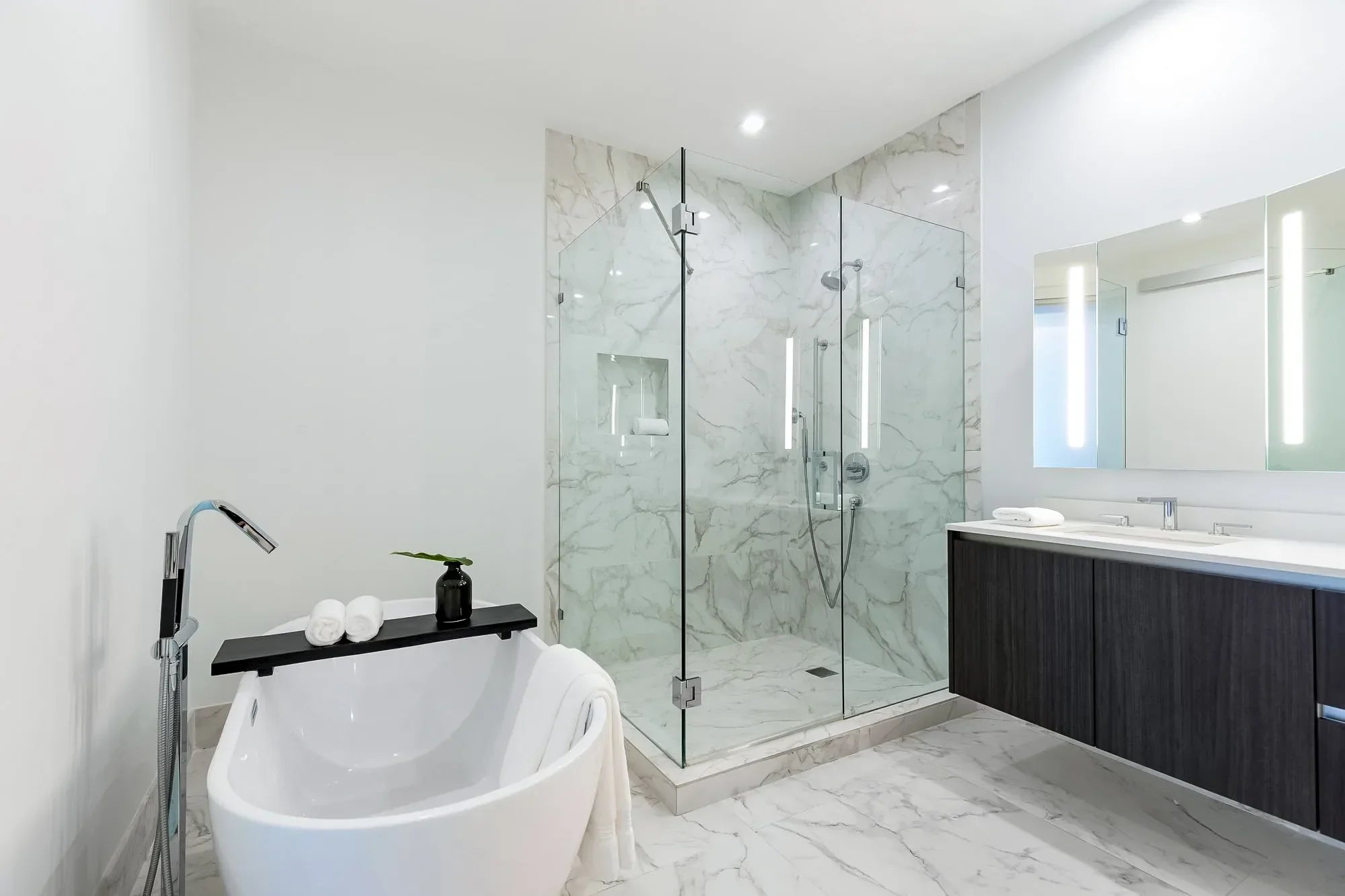 Contemporary bathroom with marble walls, frameless glass shower enclosure, freestanding soaking tub, and a floating double-sink vanity with LED mirrors.