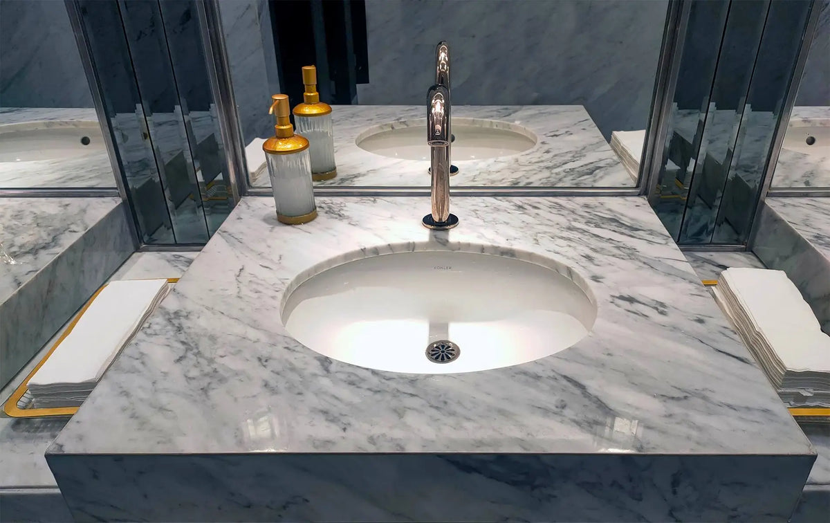 Close-up of a white Carrara marble bathroom vanity top with an undermount sink, chrome faucet, and mirrored backsplash.