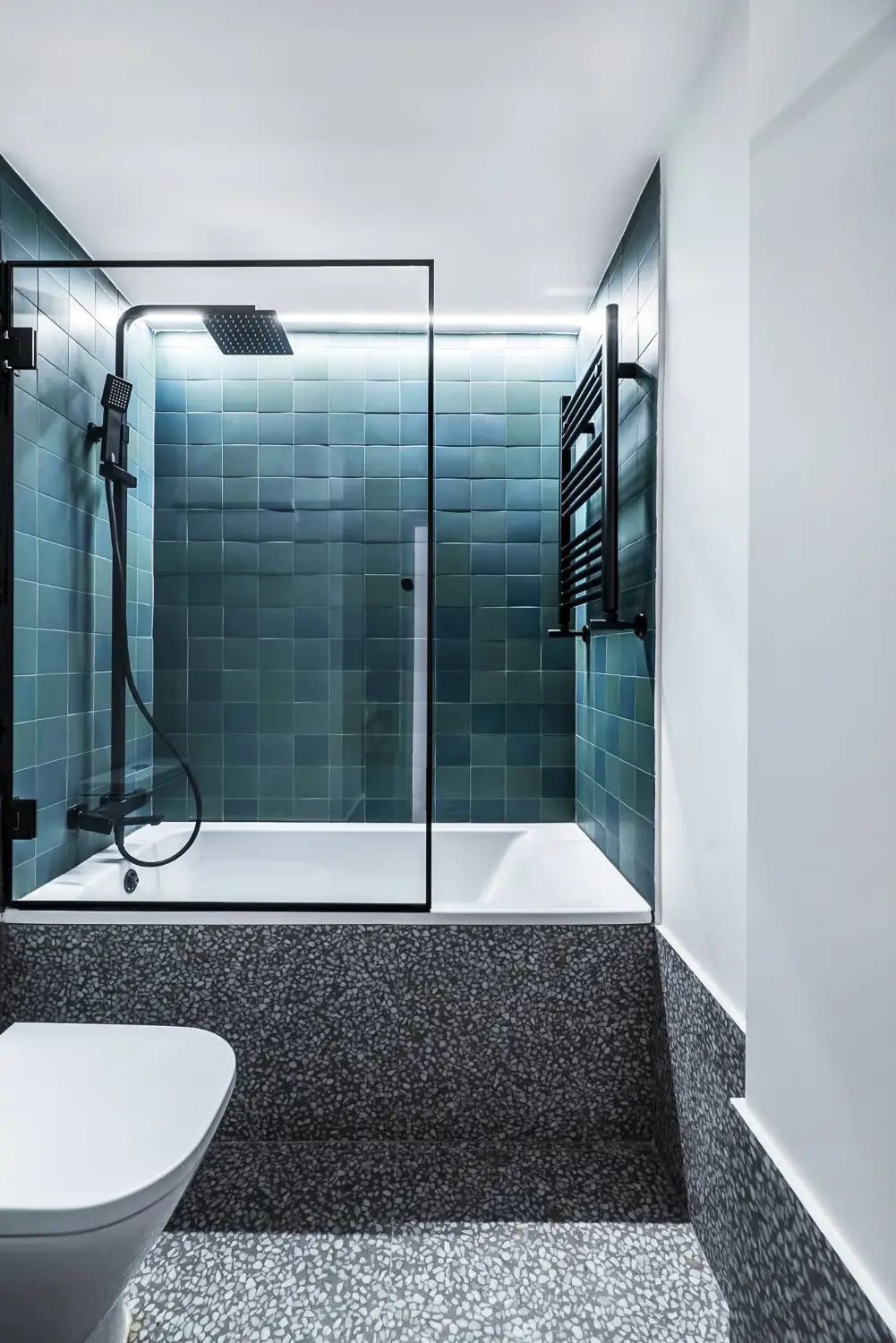 Contemporary bathroom with a bathtub and a black-framed glass shower screen, teal square tile on the shower walls, matte-black rainhead and handheld shower, terrazzo-style lower wall and floor, a wall-mounted towel warmer, recessed LED accent lighting along the tile top, and a wall-mounted toilet — a clean, modern spa-inspired design.
