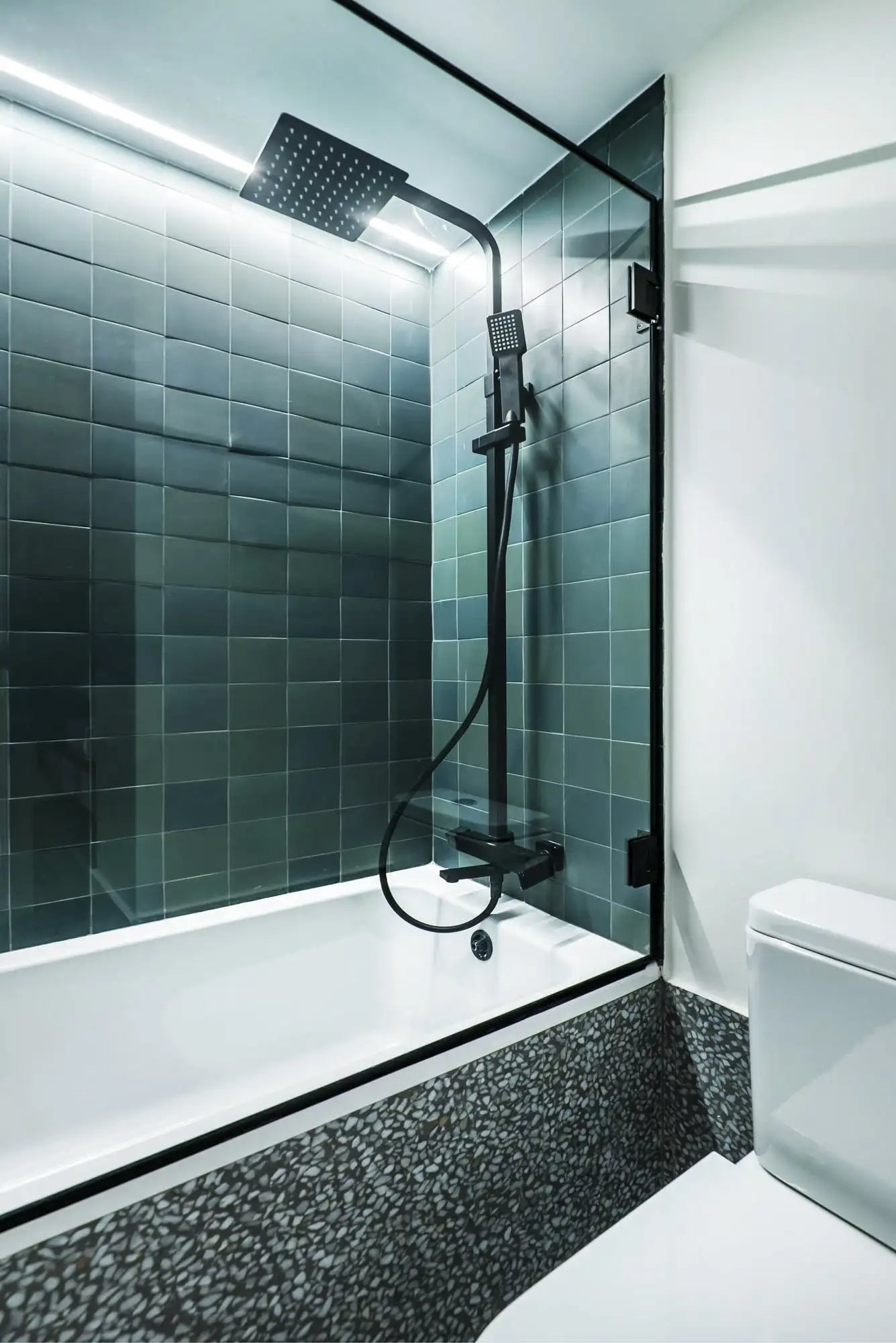 Contemporary bathroom featuring a white alcove bathtub with a black-framed glass shower screen, deep teal square tile on the shower walls, a matte-black rain shower head and handheld set, terrazzo-style tub surround and floor, recessed LED accent lighting at the tile head, and a wall-mounted toilet — a clean, modern spa-inspired design.
