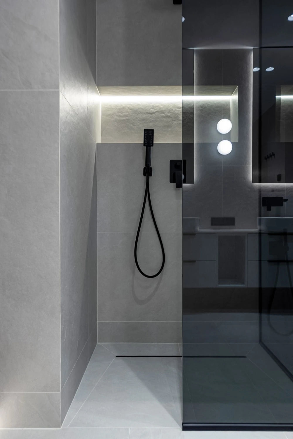 Modern walk-in shower with gray tile walls and floor, matte black handheld shower and controls, glass panel, and recessed niche with LED accent lighting.