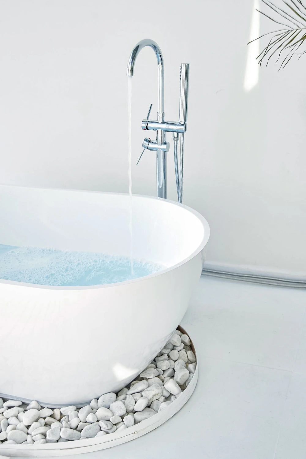 Minimalist freestanding white bathtub being filled with water, paired with a chrome floor-mounted tub filler and handheld sprayer, set against a bright white wall and surrounded by smooth white decorative stones.