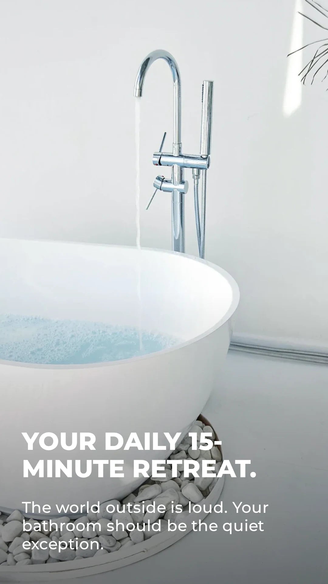 Dreamwerks modern white standalone soaking tub on a bed of white river stones featuring a floor-mounted chrome bathtub faucet with a handheld sprayer.